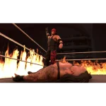 Imagen de un luchador de wrestling con ropa negra y roja de pie sobre un ring en llamas con otro luchador caído en el suelo